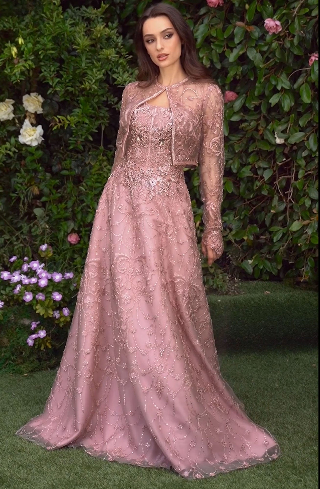 A woman stands outdoors before a green leafy backdrop, wearing the Andrea & Leo MA104 long sleeve formal dress with embroidered lace and a sheer overlay, highlighting an elegant evening style.